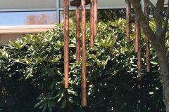 Large copper wind chimes hanging from a tree in front of a building, with lush green shrubs in the background.