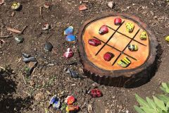 An outdoor tic-tac-toe game on a wooden tree stump slice, using hand-painted rocks decorated as ladybugs and bees as game pieces, with extra painted rocks scattered around in the dirt.