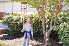 A young girl looks up at large copper wind chimes hanging from a tree in an outdoor sensory garden, with copper leaf decorations and a birdhouse also visible in the tree.