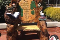 A large carved wooden storytelling chair decorated with forest animals and the text 'Once there was a beautiful forest called the Pennsylvania Wilds, and in this forest lived…' sitting on a brick patio outside a building.