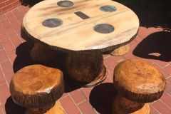 A rustic outdoor seating set made from carved tree stumps — a round table with four stump stools — sitting on a red brick patio, with four circular copper inlays and a rectangular inlay on the tabletop.