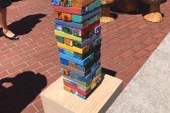 A tall outdoor Jenga-style tower game with colorful painted blocks sitting on a wooden storage box labeled 'Tumbling Tower,' on a brick patio with adults and the carved tree stump table set visible in the background.